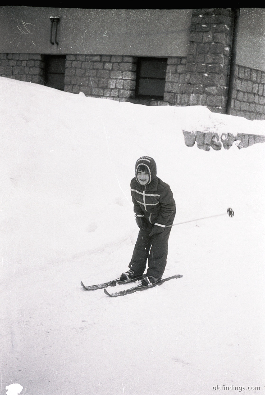 Child in vintage ski gear poses on groomed slope beside brick building, mid-20th century. Snow-covered terrain and classic ski poles suggest early skiing culture. Likely European alpine resort.