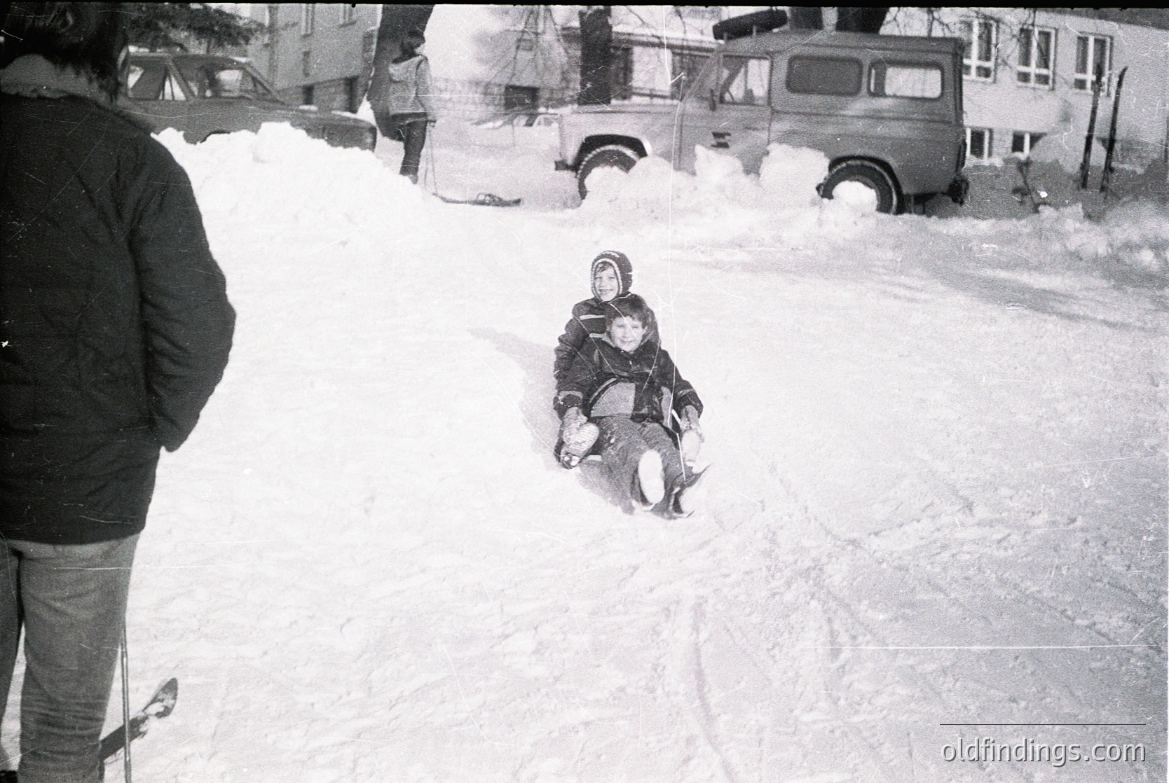 Child sledding down snow-covered street in mid-20th century urban setting. Snowdrifts obscure parked vintage vehicles and residential buildings. Photographed from behind, capturing motion and winter play.