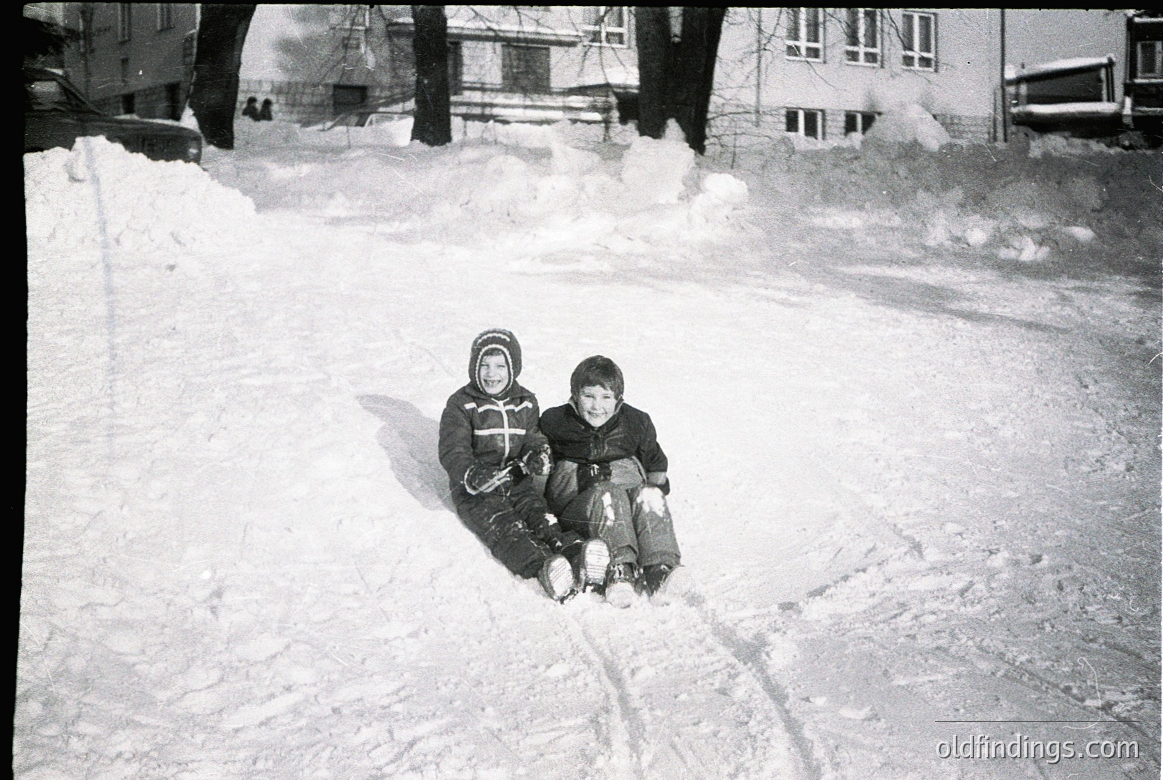 Two children sledding on a snow-covered street in mid-20th century urban setting. Snow piles and parked cars suggest residential area. Distinctive winter attire: knit caps, scarves, and heavy jackets.