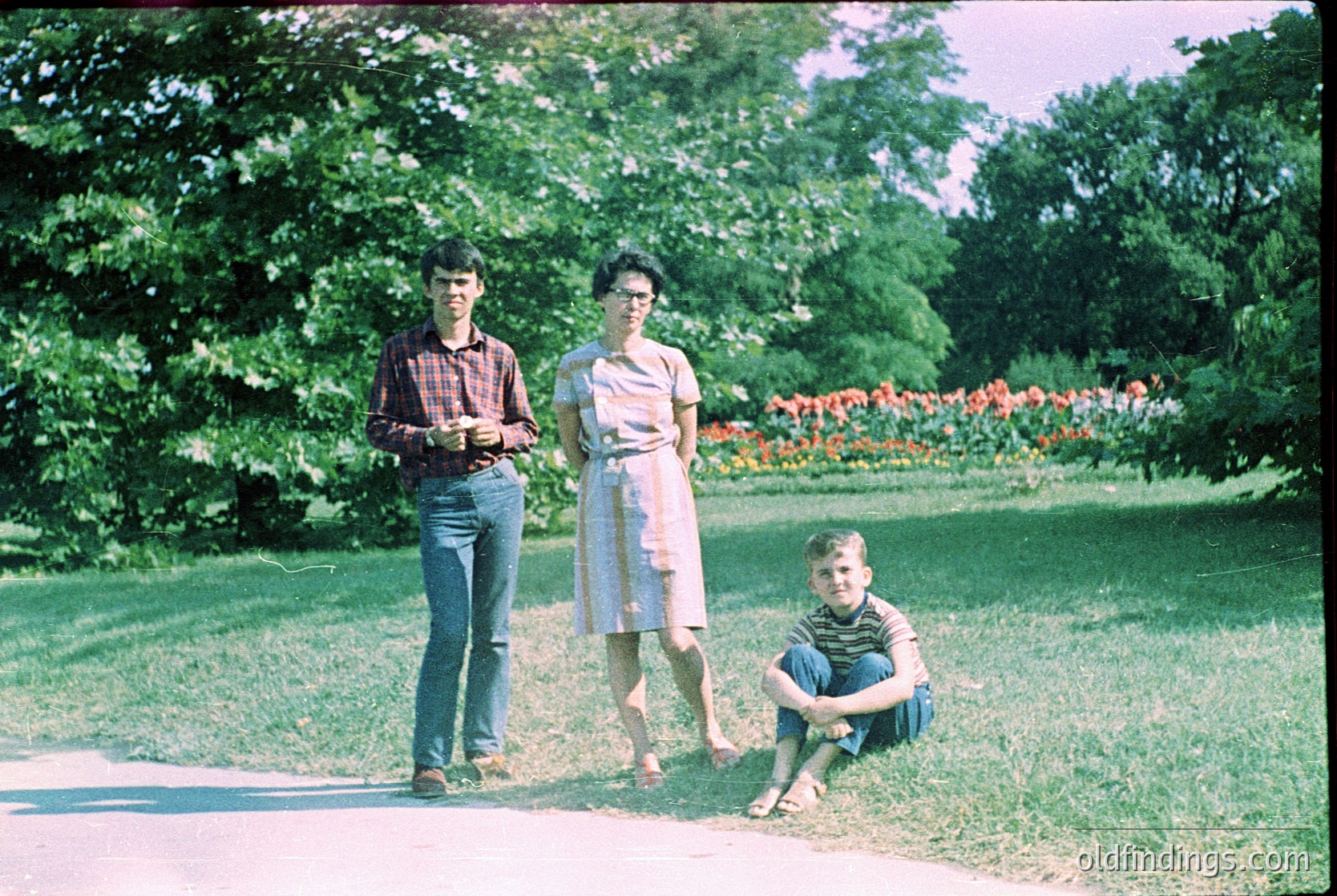 Vintage 1970s family portrait in a lush park setting. Three individuals pose on a gravel path surrounded by vibrant greenery and blooming flowers. The man wears a plaid shirt and jeans, the woman a knee-length dress with rolled sleeves, and the child a striped shirt and shorts. Colorful flowerbeds and mature trees frame the scene.