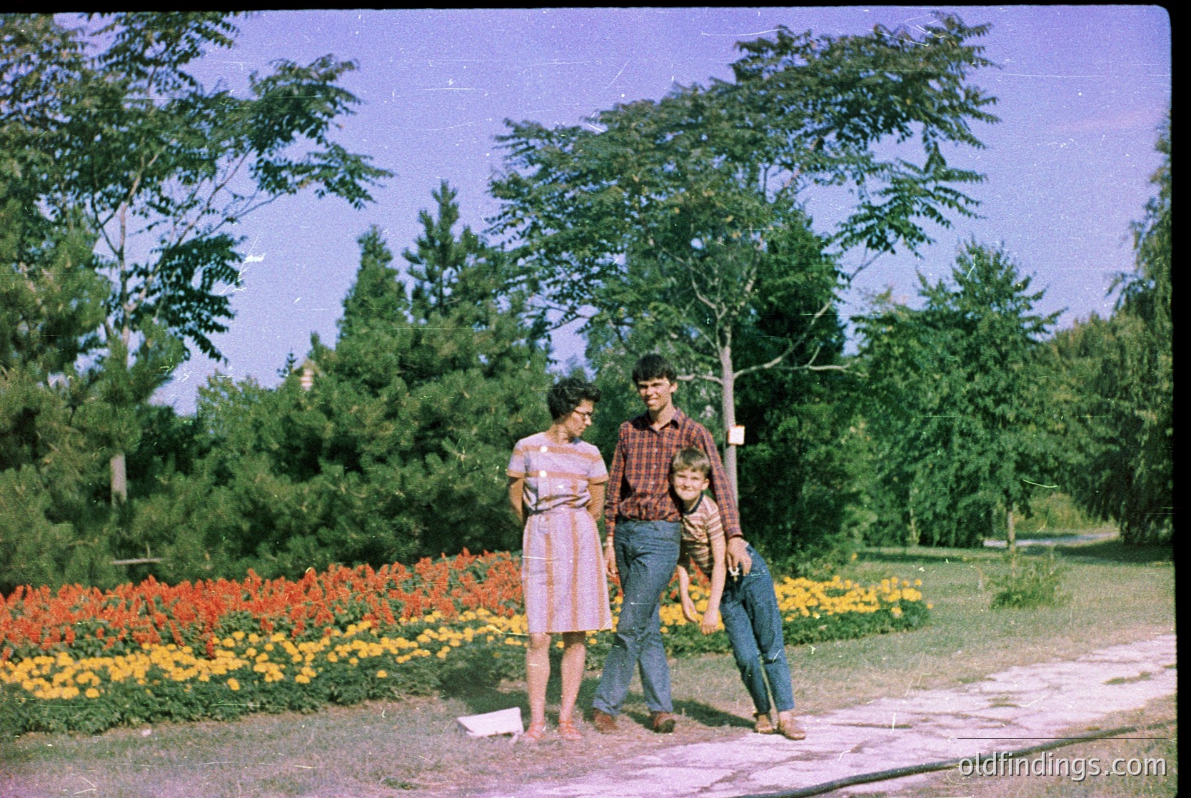 Vintage color photo of three individuals in a lush park setting, likely 1960s–1970s. Woman in floral dress, man in plaid shirt/jeans, child in shorts. Vibrant flowerbeds (orange/yellow blooms) and dense greenery frame them. Soft focus and warm tones evoke nostalgia.