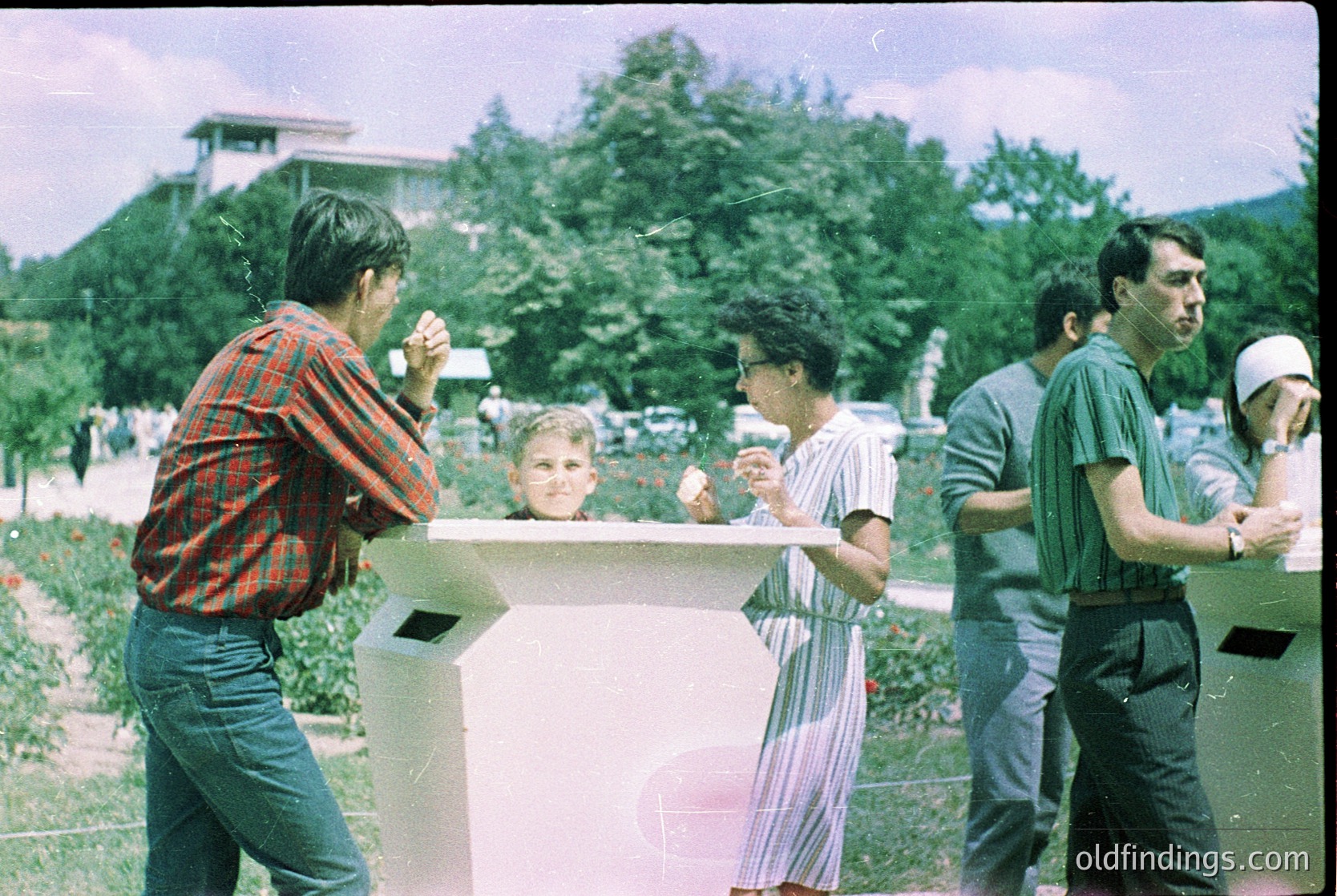 Vintage group photo in a landscaped park setting, likely 1970s–1980s. Four adults and a child pose near a concrete bench surrounded by blooming flowers. Men wear plaid shirts and caps; women in striped blouses. Lush greenery and distant buildings suggest an urban park. Ideal for nostalgic or historical research.