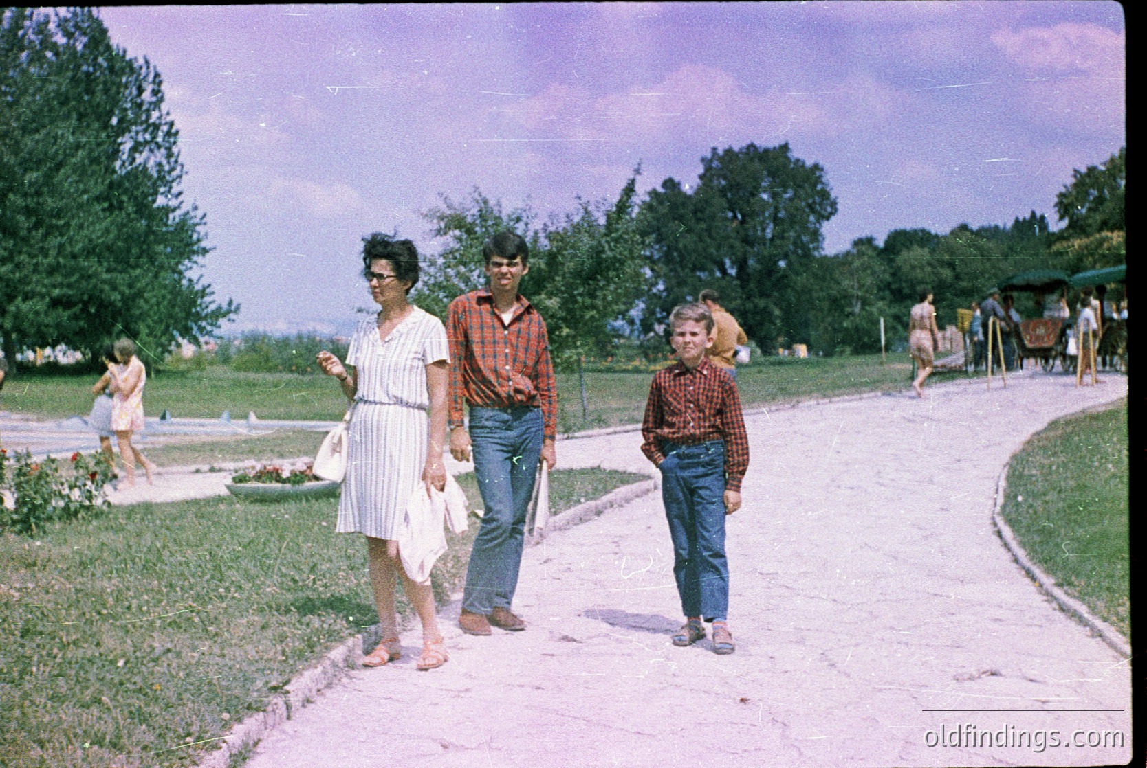 Vintage 1960s-70s park scene: woman in sleeveless dress, man in plaid shirt/jeans, and child in plaid shirt/jeans walking on paved path. Lush greenery, open sky, and distant figures in light summer attire.