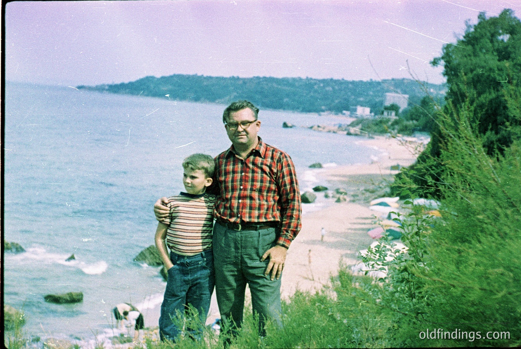 Vintage seaside portrait of a man in a plaid shirt and a young boy in striped shirt, posing on a grassy cliffside by the Black Sea coast. Mid-20th century clothing and color film suggest or . Coastal landscape includes rocky shore, waves, and distant buildings.