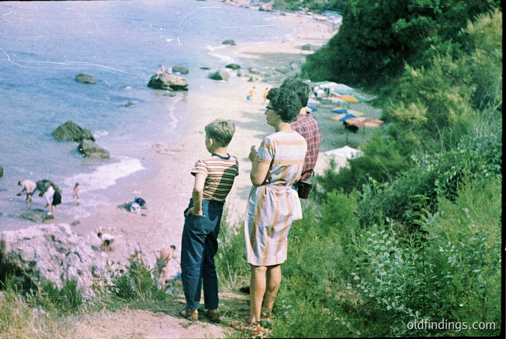 A mid-20th century seaside scene: two adults and a child stand on a grassy cliff overlooking a rocky shoreline. The woman wears a striped dress and glasses; the boy sports a striped shirt and shorts. In the background, beachgoers swim and sunbathe near colorful umbrellas. Vibrant vintage filter suggests 1950s–1960s coastal leisure.
