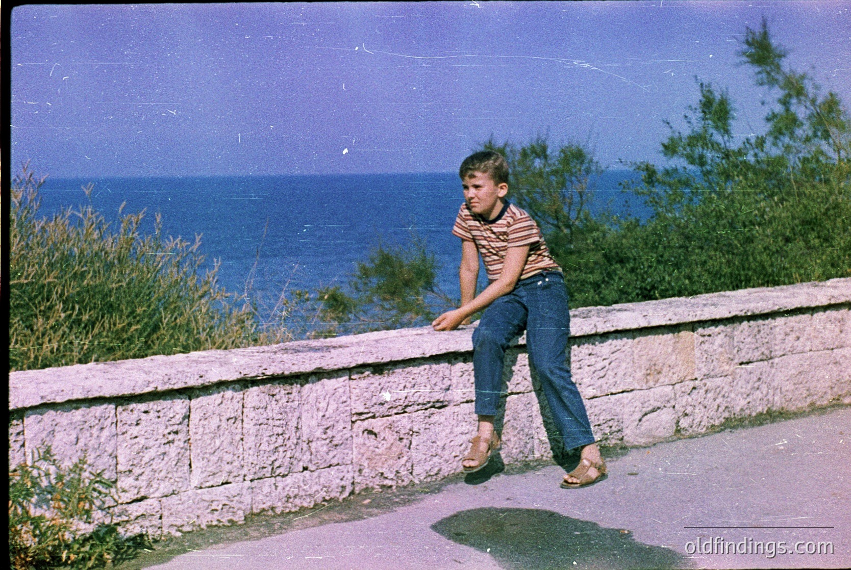 Vintage seaside shot of a boy in 1960s-style striped shirt and jeans leaning on a stone wall, overlooking the ocean. Warm sepia tone captures mid-century coastal vibes.