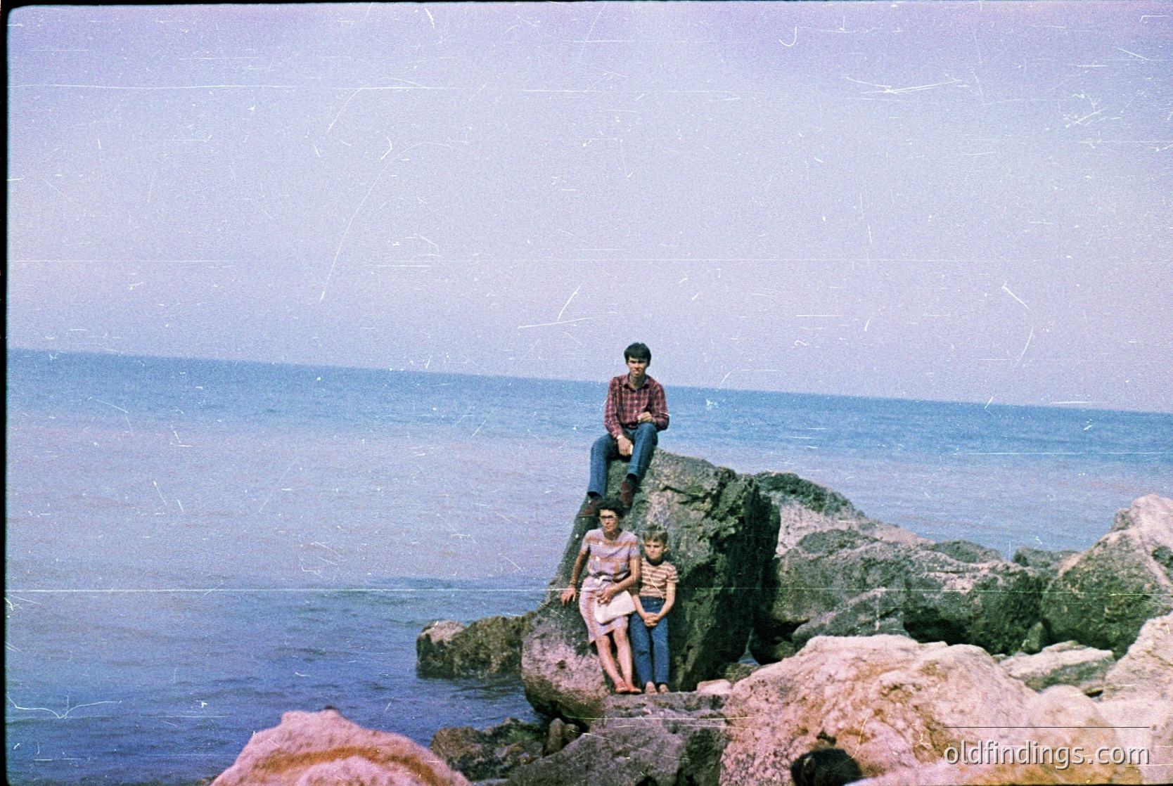 Vintage seaside family portrait on rugged rocks, likely Mediterranean. Three individuals pose on uneven stone formations near turquoise waters, suggesting a mid-20th-century beach outing. The man wears a plaid shirt, woman a floral dress, and child a light-colored top. Faded film grain and color saturation hint at 1960s-1970s photography.