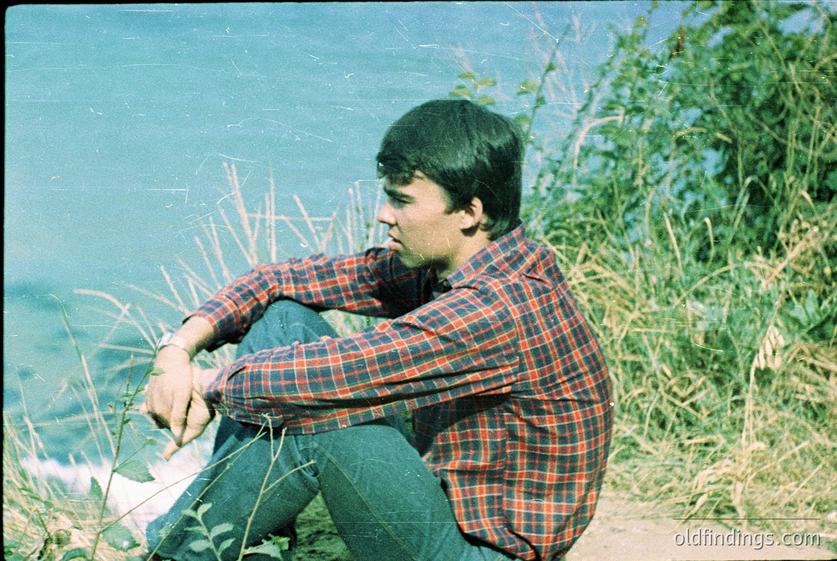 Young man in 1970s plaid shirt sits contemplatively by a coastal shoreline, hands clasped. Vintage sepia-toned photo captures natural light and grassy foreground. Reflective water and distant horizon suggest a serene seaside setting.