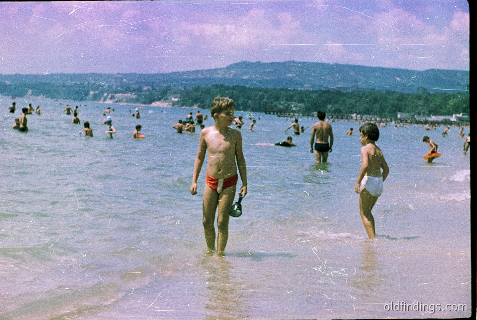 Vintage seaside scene with two boys in shallow water, one wearing red shorts, the other white. Crowded beach with adults and children swimming, framed by distant forested hills. Color-tinted photo suggests 1950s–1960s era.