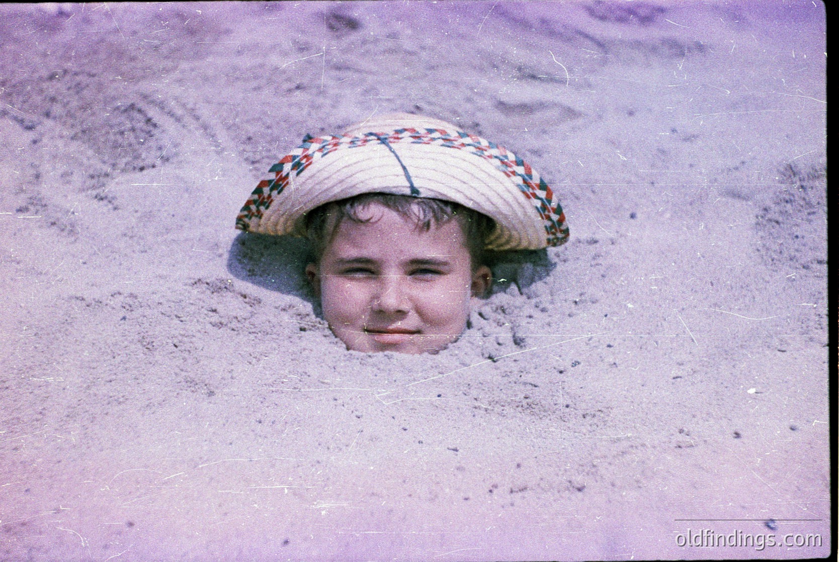 Child peeking from a sand tunnel, wearing a wide-brimmed straw hat with colorful woven trim. Mid-20th century beach scene, likely Eastern European coastal area. Warm sepia-toned vintage photograph.