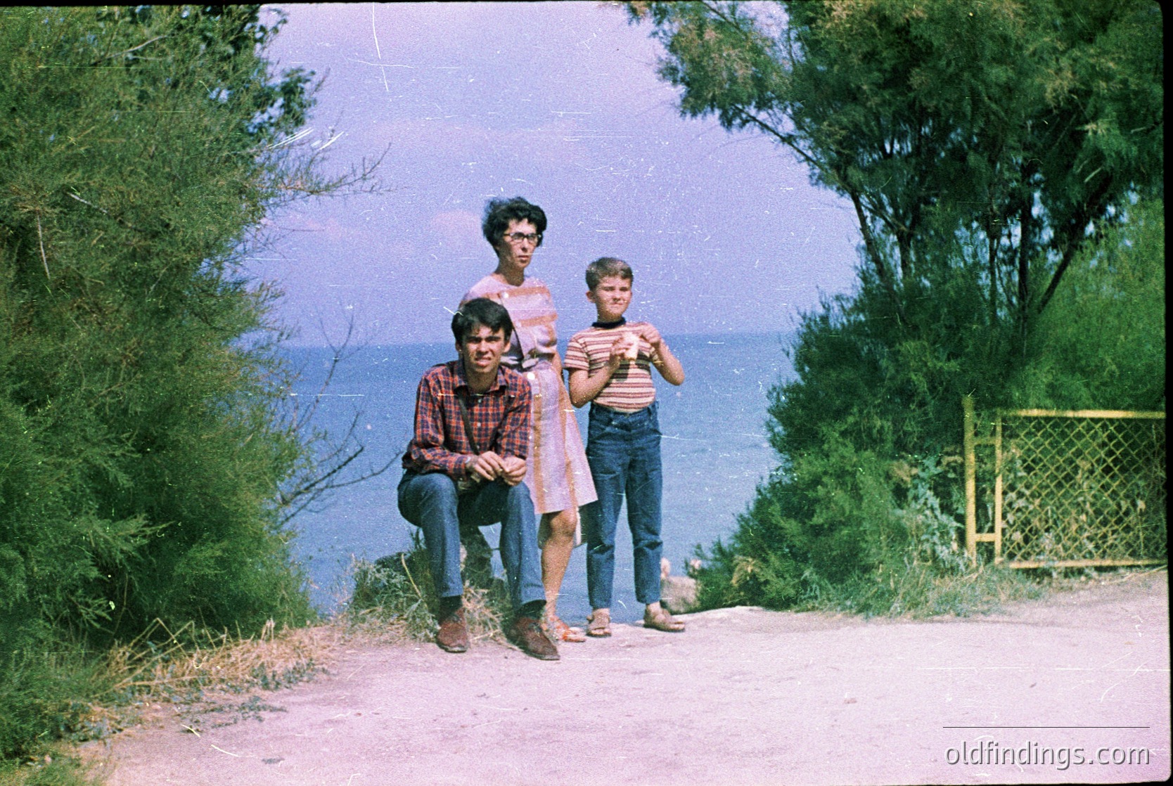 Family portrait by a seaside cliff, 1970s. Three individuals pose on a gravel path with ocean backdrop, framed by greenery. The woman wears a floral blouse and dark skirt; the boy on left sports a plaid shirt and jeans, the boy on right a striped shirt and overalls. Vintage color film captures nostalgic coastal vibes.
