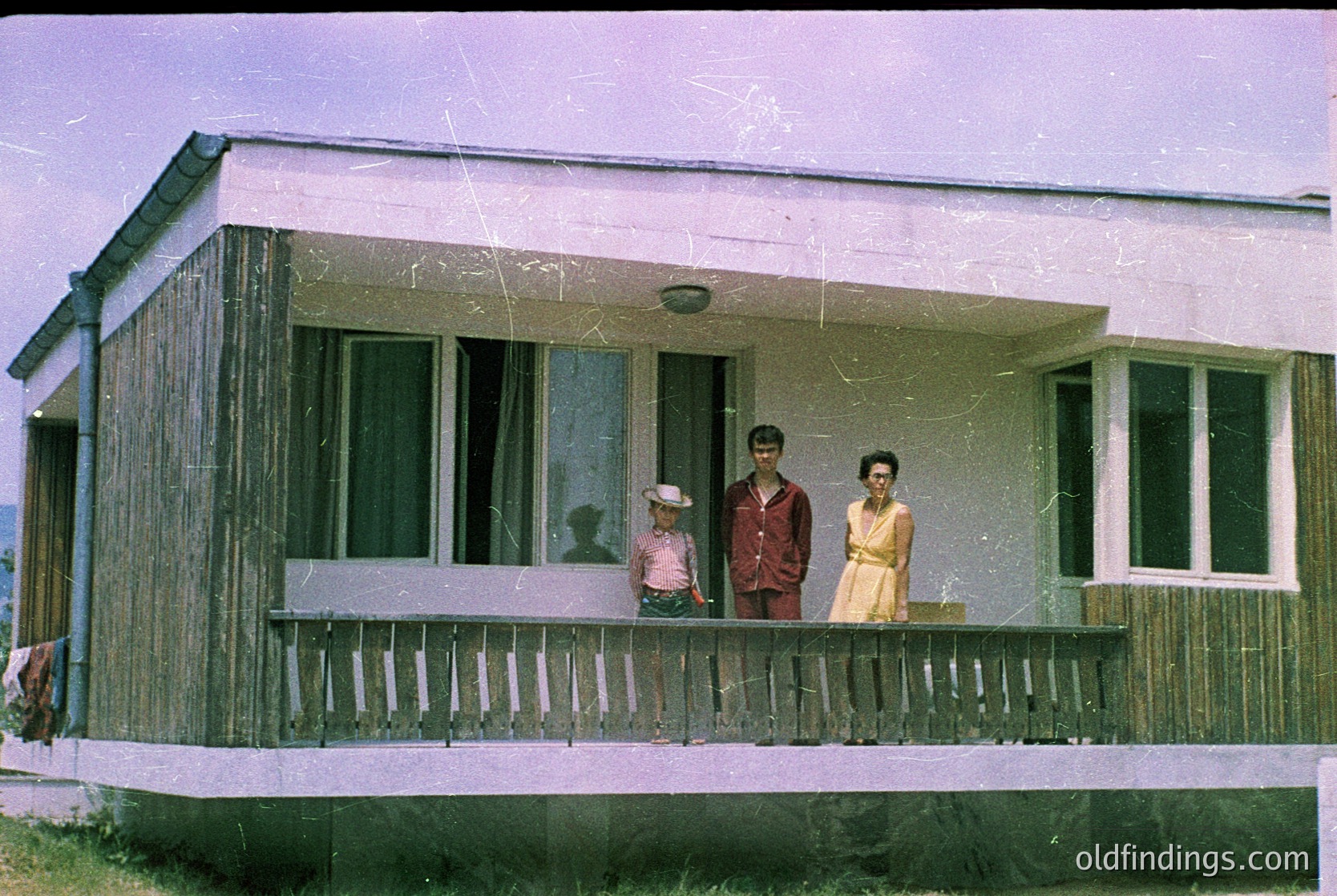 Mid-century prefab home with wooden paneling and wide porch railings. Three adults pose on balcony: man in dark jacket, woman in light dress, child in hat. Vintage sepia-toned photo suggests 1950s–1960s era. Rural or suburban setting with minimal background detail.