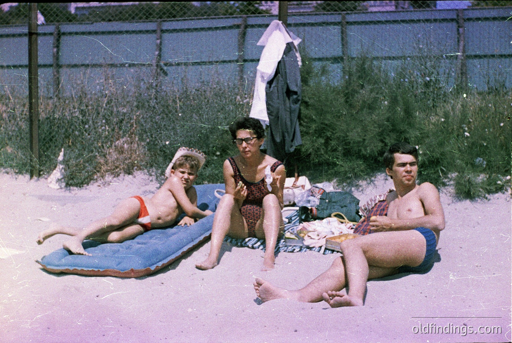Family beach scene from the 1960s–70s, featuring three generations lounging on a faded towel. Adults in swimwear and glasses, child in red-and-white striped swimsuit. Wooden fence and beachside vegetation in background. Authentic vintage summer vibes.