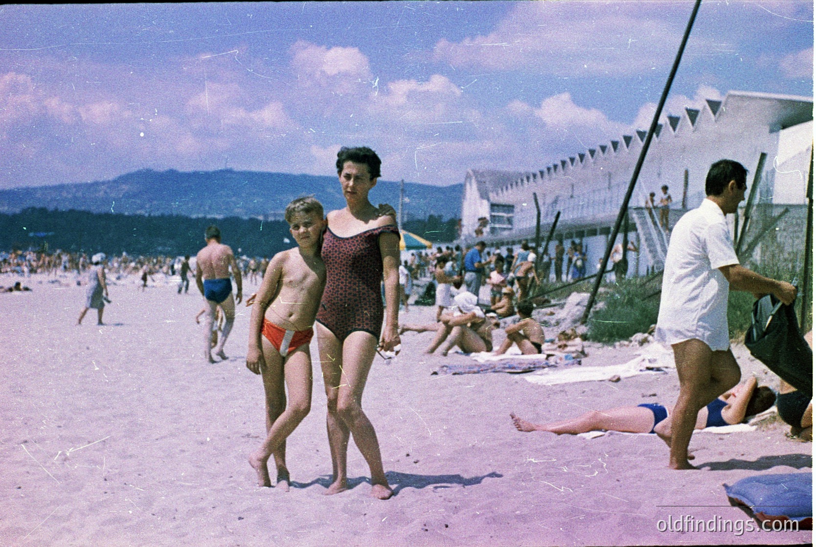 Vintage seaside scene from mid-20th century, likely 1950s–1960s. A woman in a sleeveless dress and a young girl in red-and-white striped one-piece swimwear pose on a sandy beach. Crowds relax under striped beach umbrellas, while lifeguard towers and mountains frame the horizon. Colorful, nostalgic beach culture.