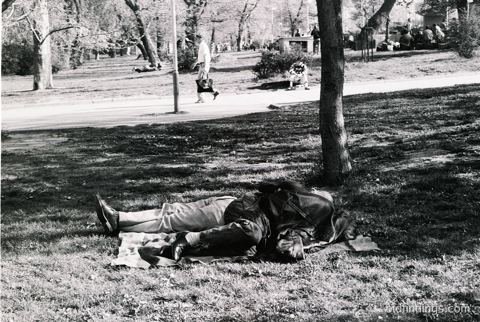 Black-and-white park scene from the 1960s–70s: two individuals lying on grass beneath mature trees, one reading a book, the other resting with legs elevated. Mid-century urban park design with paved paths and distant park-goers. Evokes mid-20th-century leisure culture.
