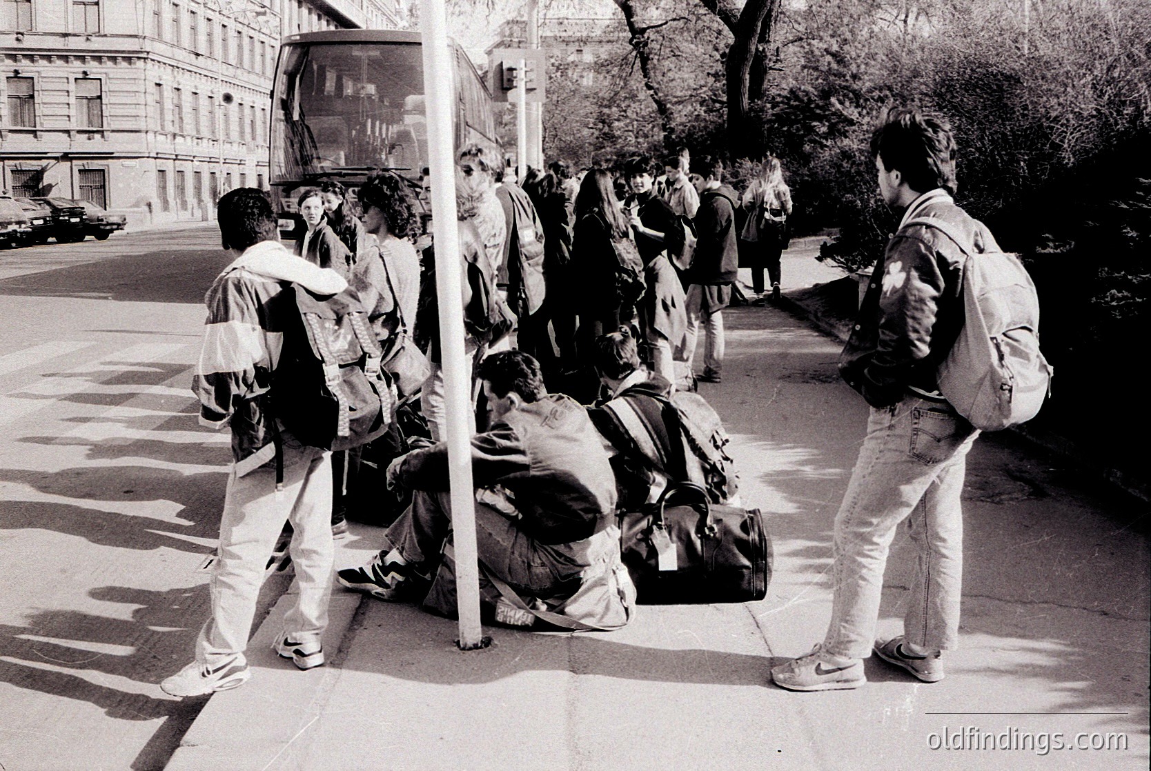 Black-and-white street scene featuring a group of young people waiting at a bus stop in an urban setting. One person sits on the curb with a large backpack, while others stand or lean against a pole. A vintage bus shelter and classic cars in the background suggest a mid-20th-century European city. Casual, youthful attire and relaxed postures indicate a moment of transit or socializing.