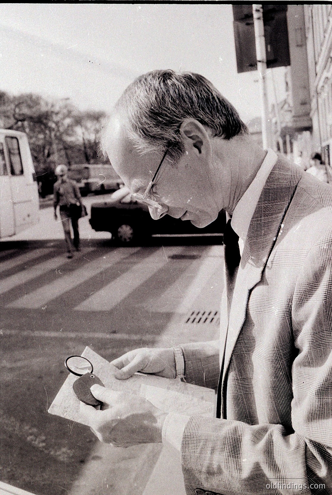 Mid-20th century man examines a map with a magnifying glass on a city street, likely 1950s–1960s. Urban setting with parked vehicles and pedestrians in the background. Formal attire suggests professional or official activity.