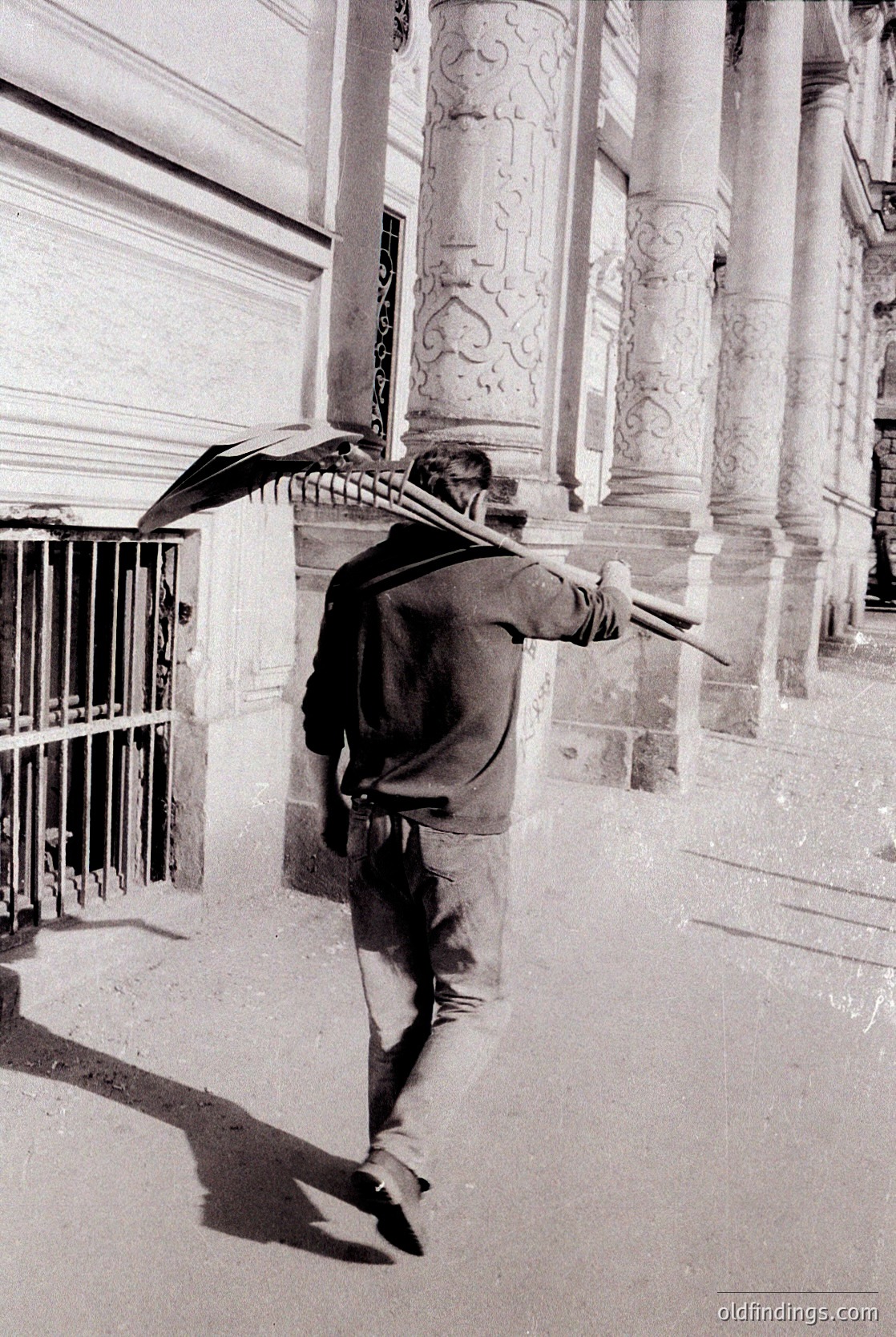 Mid-20th century street scene: Man in mid-stride under ornate umbrella, holding a second open umbrella. Architectural details include fluted columns with decorative capitals and wrought-iron railings. Likely urban European setting, possibly 1950s–1960s.