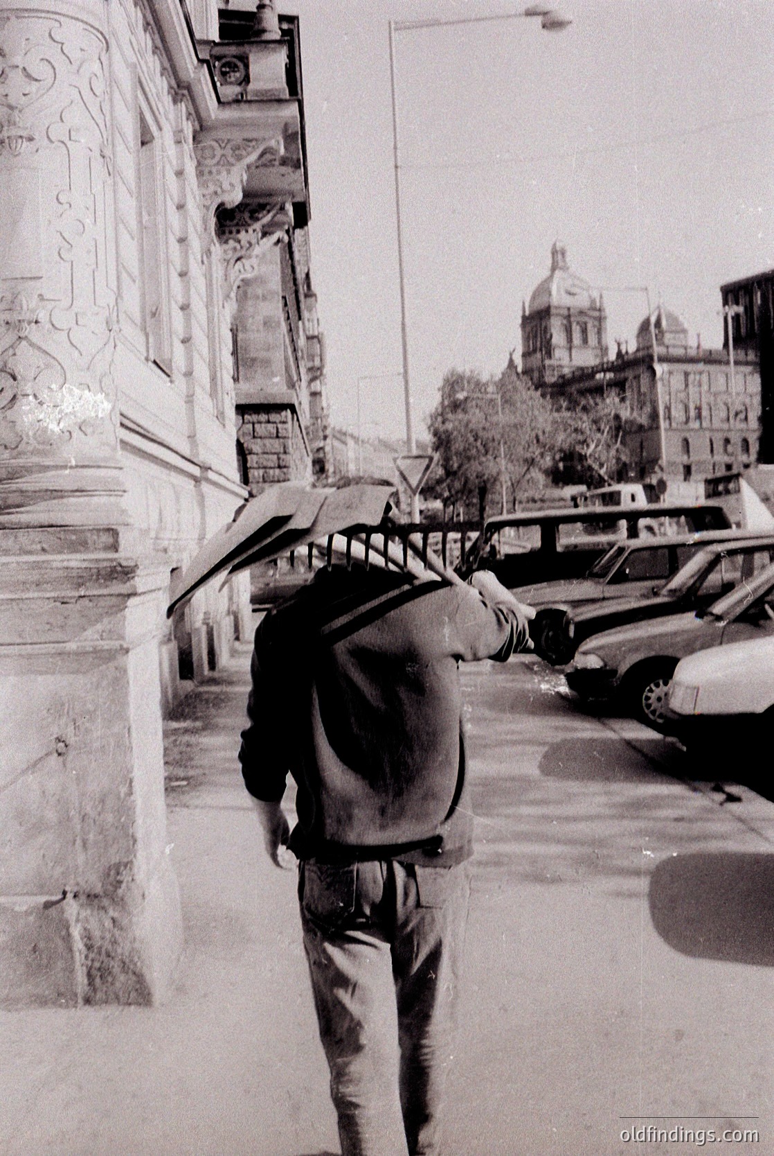 Man in 1970s-style jacket and backpack leans against ornate stone pillar, adjusting a folded umbrella. Urban street scene with parked cars, Soviet-era architecture, and distant domed building. Likely Eastern Europe.