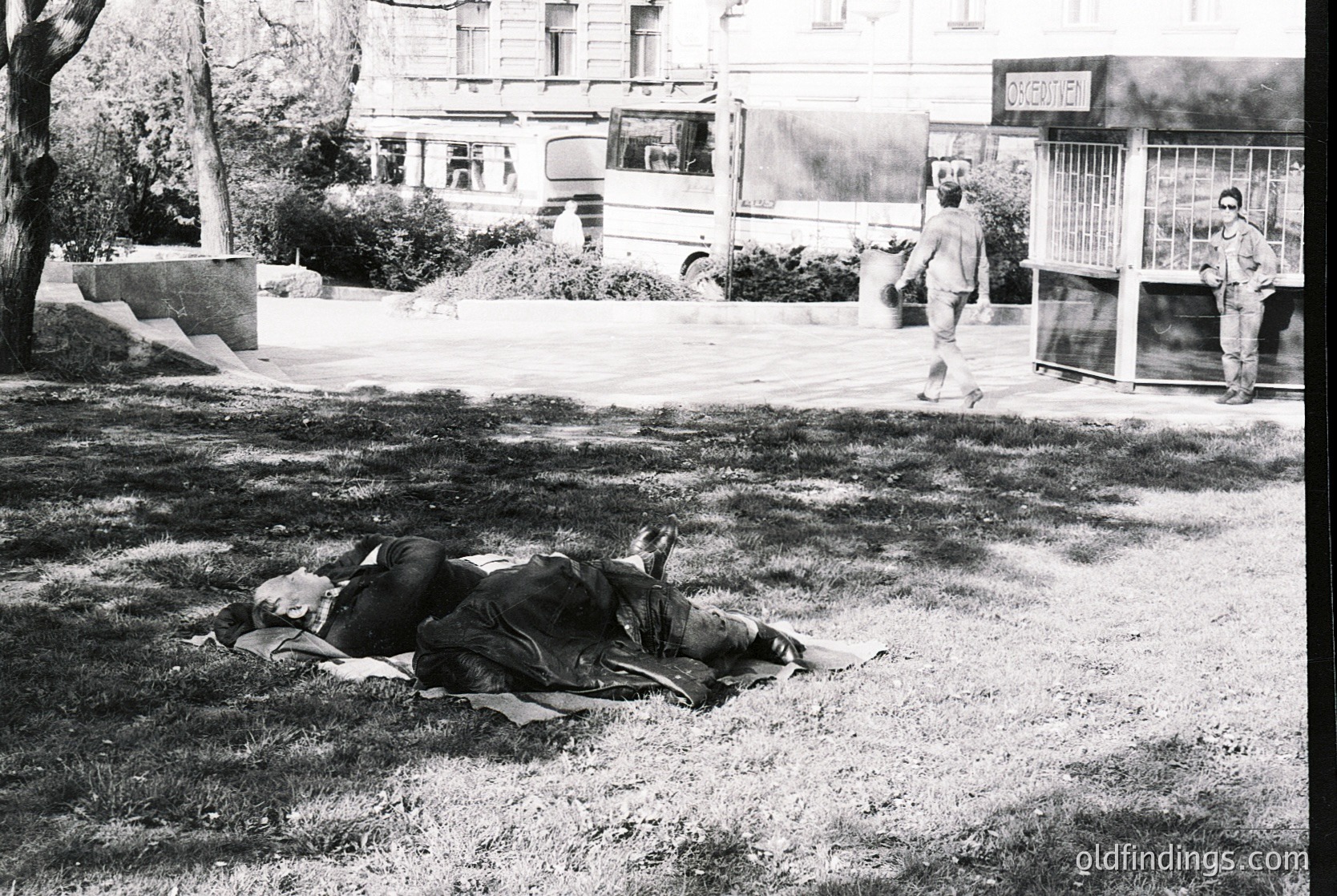 Black-and-white street scene showing two individuals lying prone on grass, likely during a protest or demonstration. Visible signage reads "EXPROPRIEREN" (Dutch/German for "expropriate") and "BUSSENSTATION" (Dutch for "bus station"). Urban setting with parked buses, trees, and a bus shelter. Mid-20th century European city atmosphere.