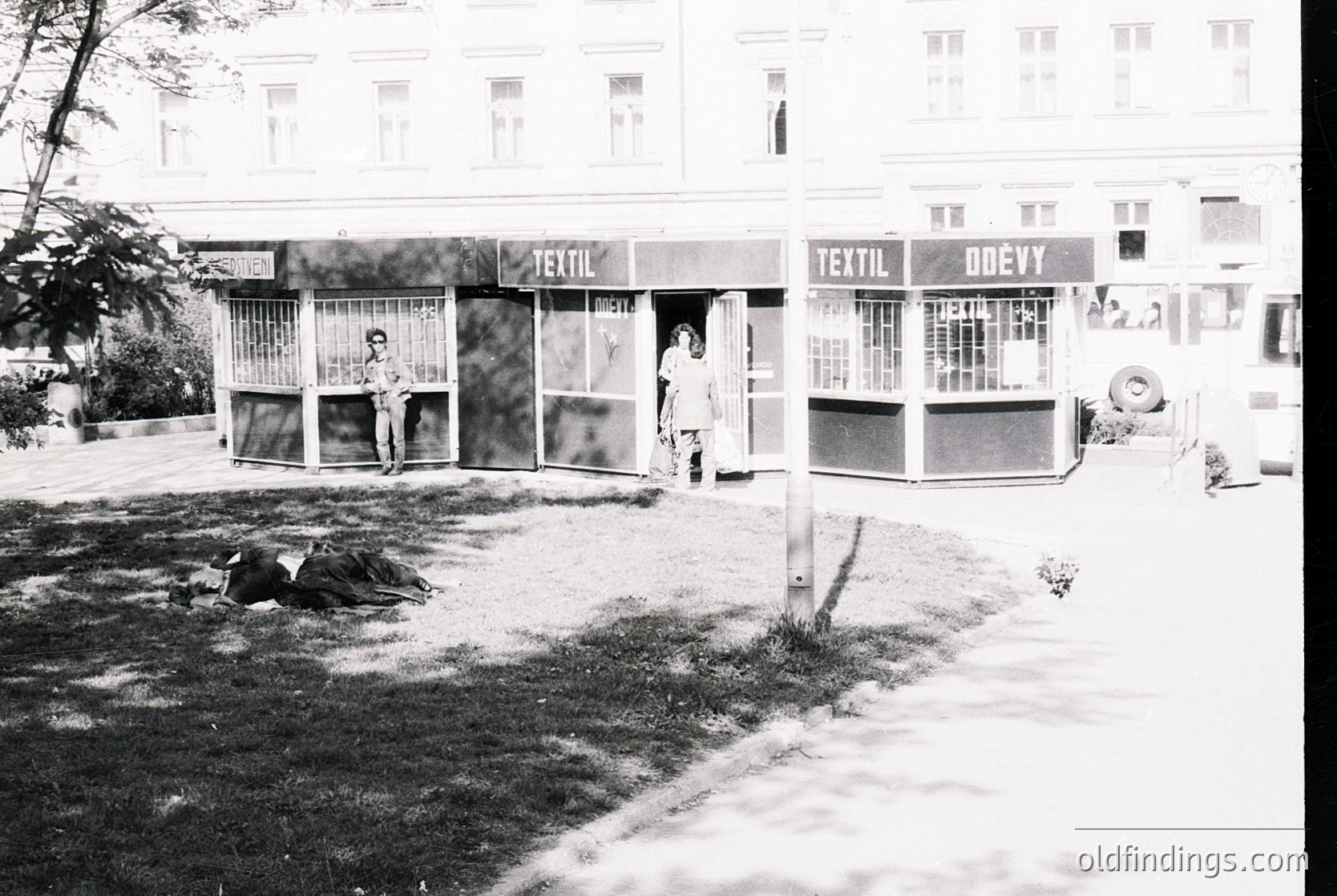 Black-and-white street scene featuring two textile shops with signage "Textil" and "Textil Onev" in Eastern Bloc architectural style. Two men in casual 1960s-70s attire stand near open shop fronts, while a third reclines on grass. Urban setting with bare trees and paved sidewalk.