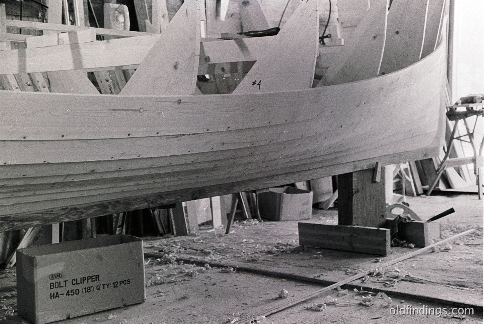 Mid-20th century boat construction in progress, featuring plywood hull assembly with visible rivets and clamps. Box labeled "Bolt Clipper HA-450 (18)" indicates marine hardware. Workshop setting with scattered wood shavings and tools.