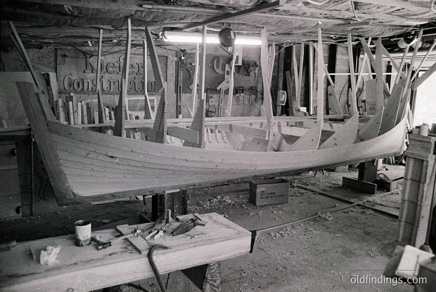Traditional wooden boat under construction in a workshop, framed ribs visible. Sign reads "Kiel Construction." Mid-20th century craftsmanship with hand tools and unfinished hull.