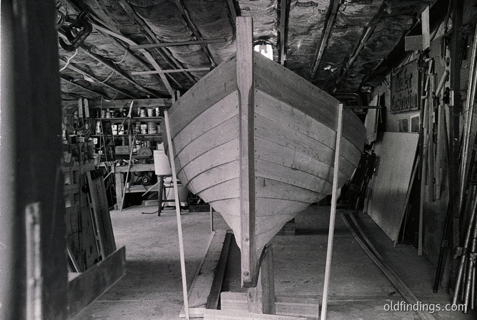 Mid-20th century workshop showcasing traditional wooden boat construction. Planks and ribs form the hull of a small vessel, supported by metal braces. Shelves and tools line the walls, indicating a functional crafting space. Black-and-white format suggests archival or historical significance.