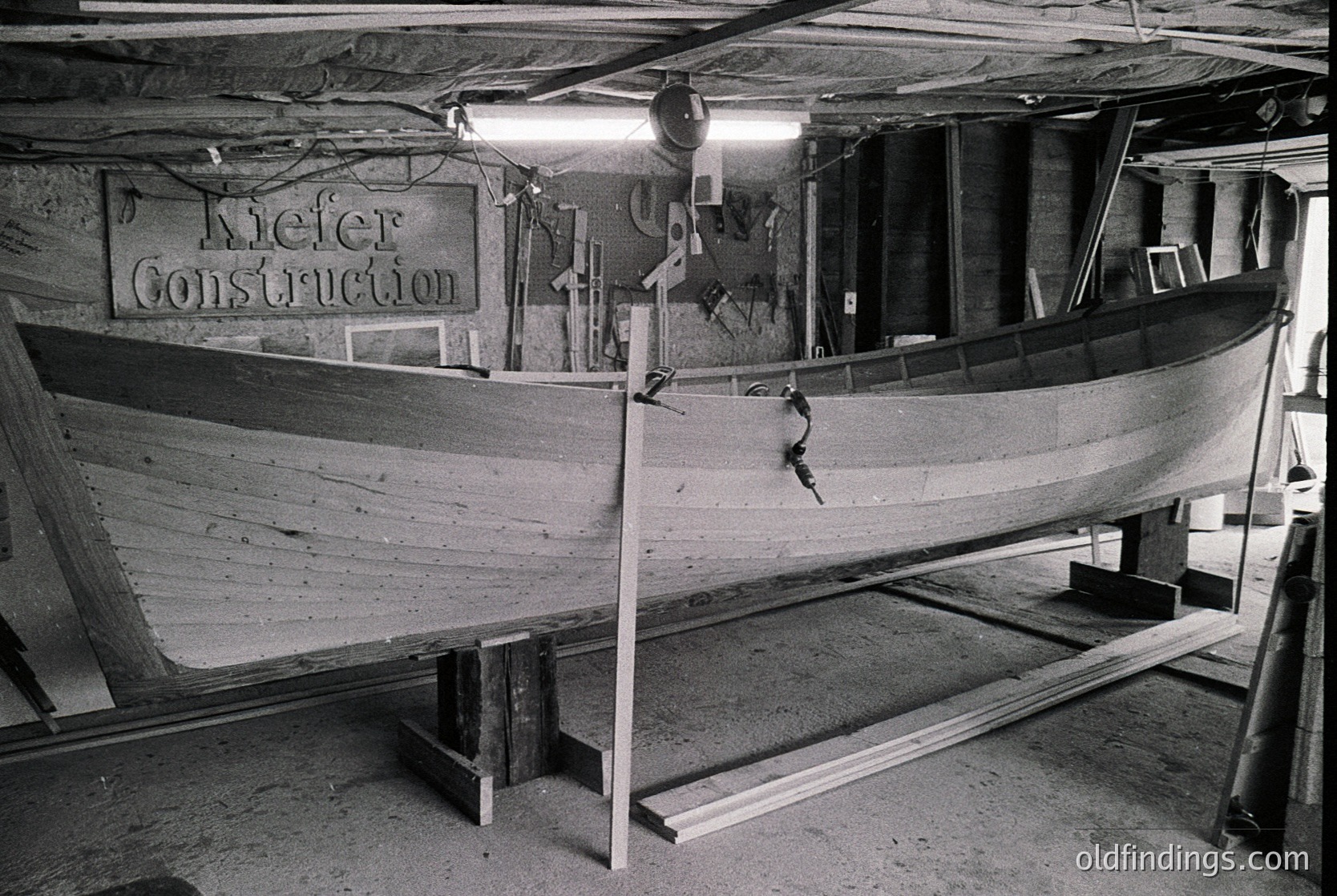 Industrial boat construction scene inside a workshop labeled "Kiefer Construction." A wooden boat hull rests on supports, with visible rivets and structural reinforcements. Overhead lighting and exposed pipes indicate mid-20th century workshop design.