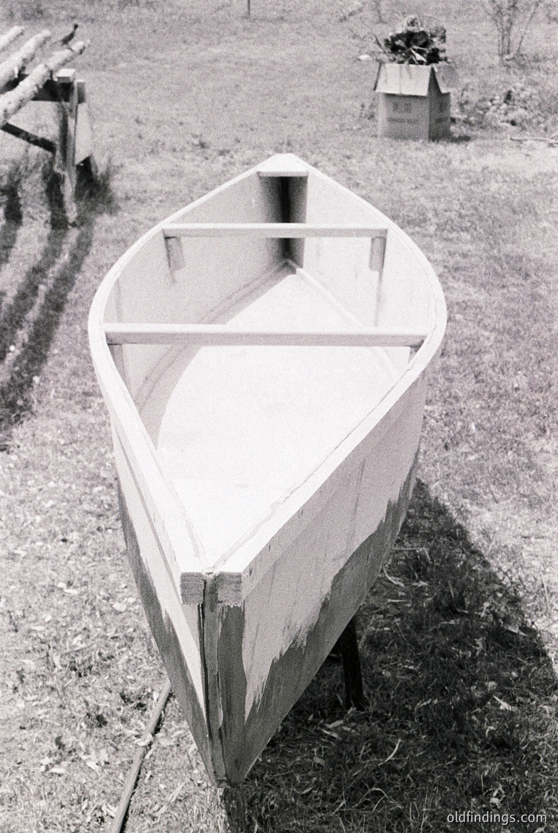 Classic wooden rowboat with simple frame structure, resting on grassy ground near a dirt path. Rustic wooden fence and small shed in background. Likely mid-20th century rural setting.