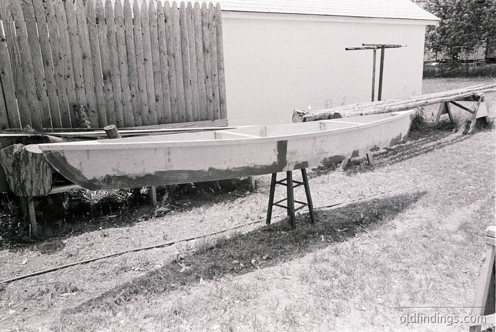 Vintage wooden boat resting on a makeshift cradle beside a weathered wooden fence and small shed. Rustic rural setting, likely early-to-mid 20th century.