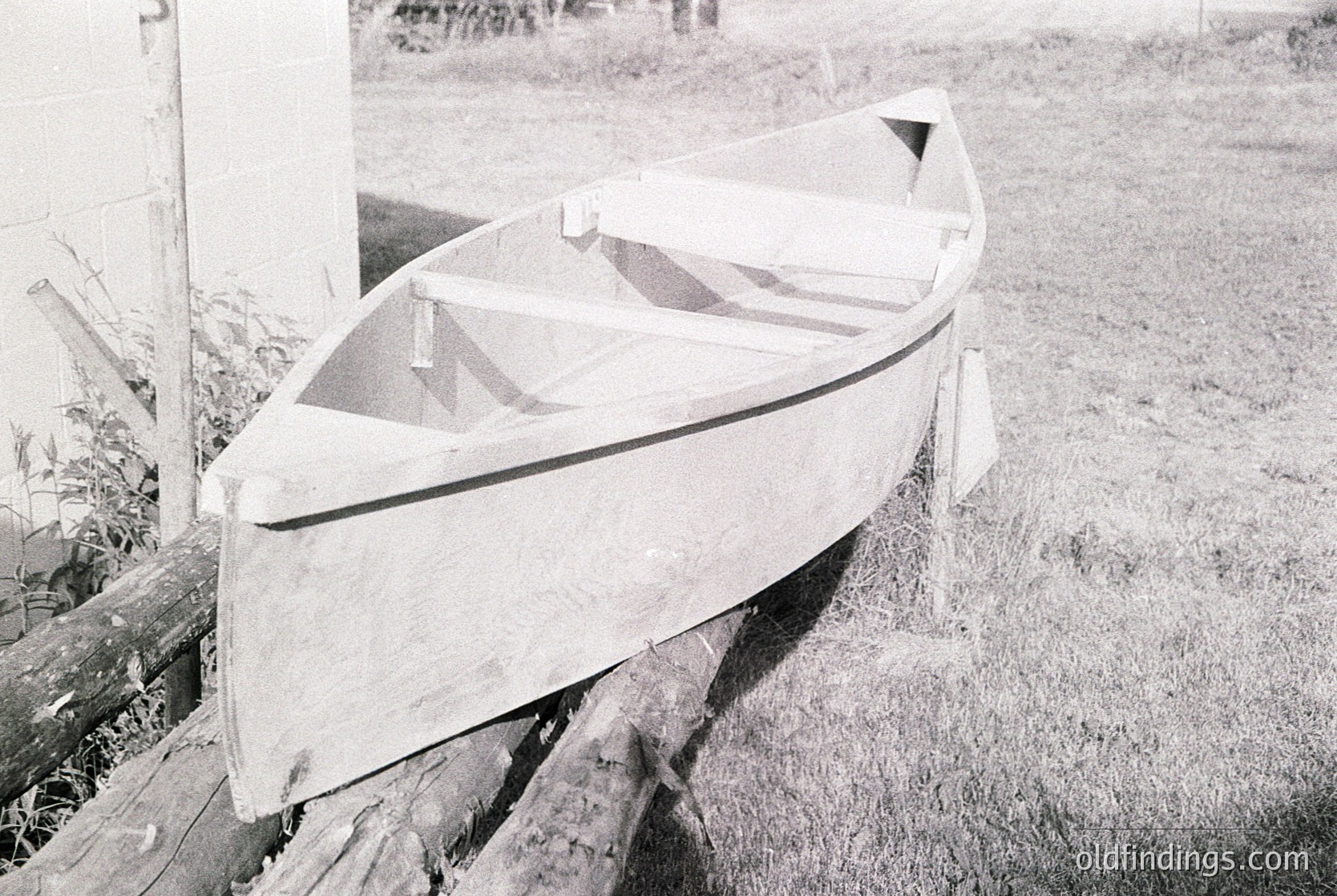 Classic wooden rowboat resting on wooden supports, likely mid-20th century. Smooth hull with simple, functional design, indicative of traditional craftsmanship. Grass and overgrown vegetation suggest rural or lakeside setting.