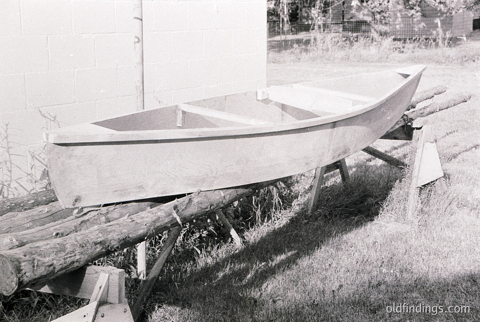 Vintage wooden boat resting on makeshift supports in a grassy area, likely mid-construction or repair. Visible planks, nails, and a simple frame suggest traditional craftsmanship. Background shows a fence and overgrown vegetation, indicating rural or coastal setting. Black-and-white suggests 20th-century origin.