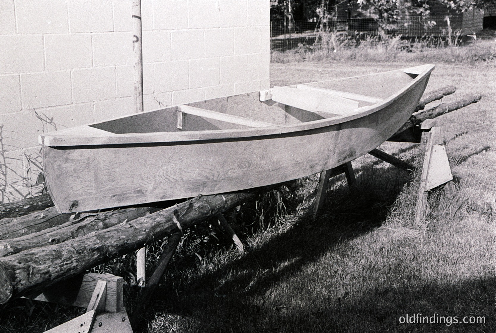 Handcrafted wooden boat on wooden supports, likely mid-construction or repair. Plain hull with minimal detailing, resting on a grassy area beside a brick wall. Black-and-white monochrome suggests vintage or archival quality. Potential 20th-century industrial or maritime setting.