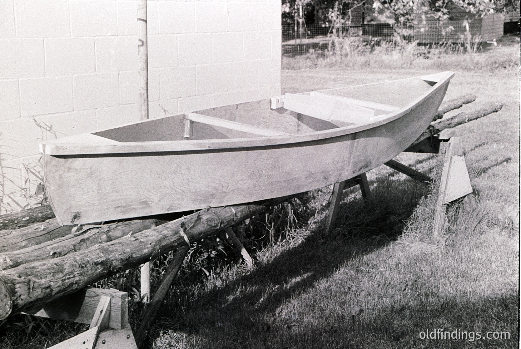 Classic wooden rowboat resting on makeshift wooden supports in a grassy yard. Mid-20th century construction with smooth, rounded hull and simple design.