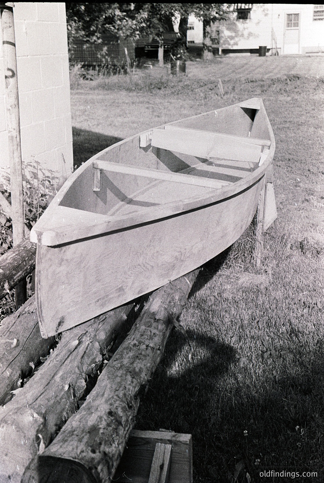 Classic wooden dinghy resting on a wooden cradle, likely mid-20th century. Simple, functional design with a flat bottom and minimalist lines, suggesting coastal or lakeside use. Background shows a brick wall and overgrown grass, hinting at a rural or semi-rural setting.