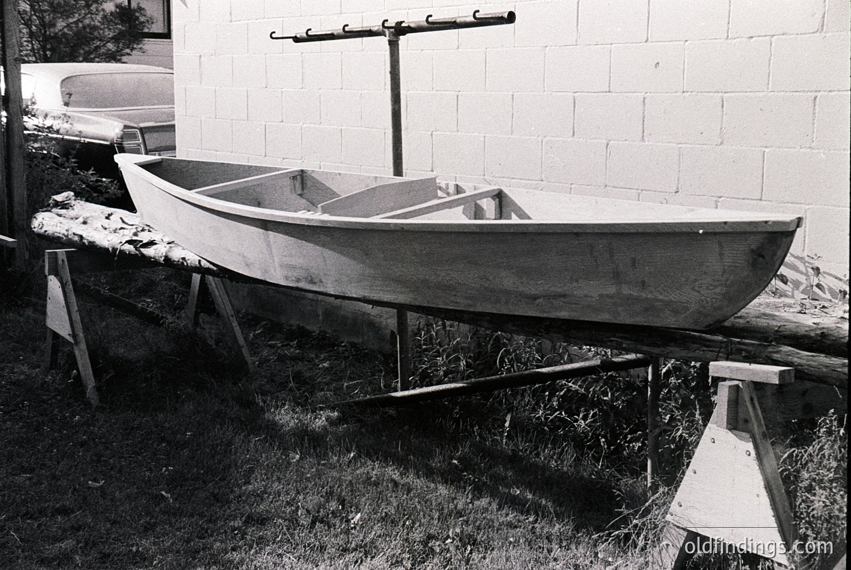 Handcrafted wooden boat on elevated wooden supports, likely a fishing or utility vessel. Simple, functional design with a flat bottom and minimalist construction. Partial view of a vintage car in background suggests mid-20th century setting.