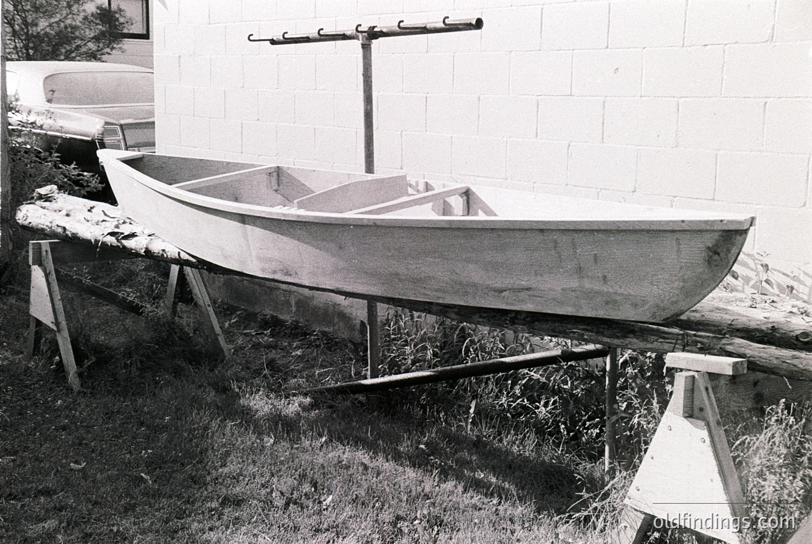 Classic wooden rowboat elevated on supports, likely mid-20th century. Simple, functional design with a single bench seat and outrigger stabilizer. Positioned outdoors near a brick wall, suggesting a coastal or lakeside setting. Ideal for vintage maritime or historical research.