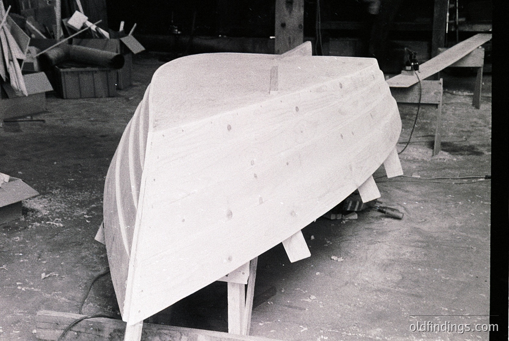 Mid-century industrial boat hull in fiberglass construction, draped in protective cover. Factory setting with wooden molds, tools, and unfinished materials. Likely 1950s–1970s era, reflecting post-war boatbuilding innovation.