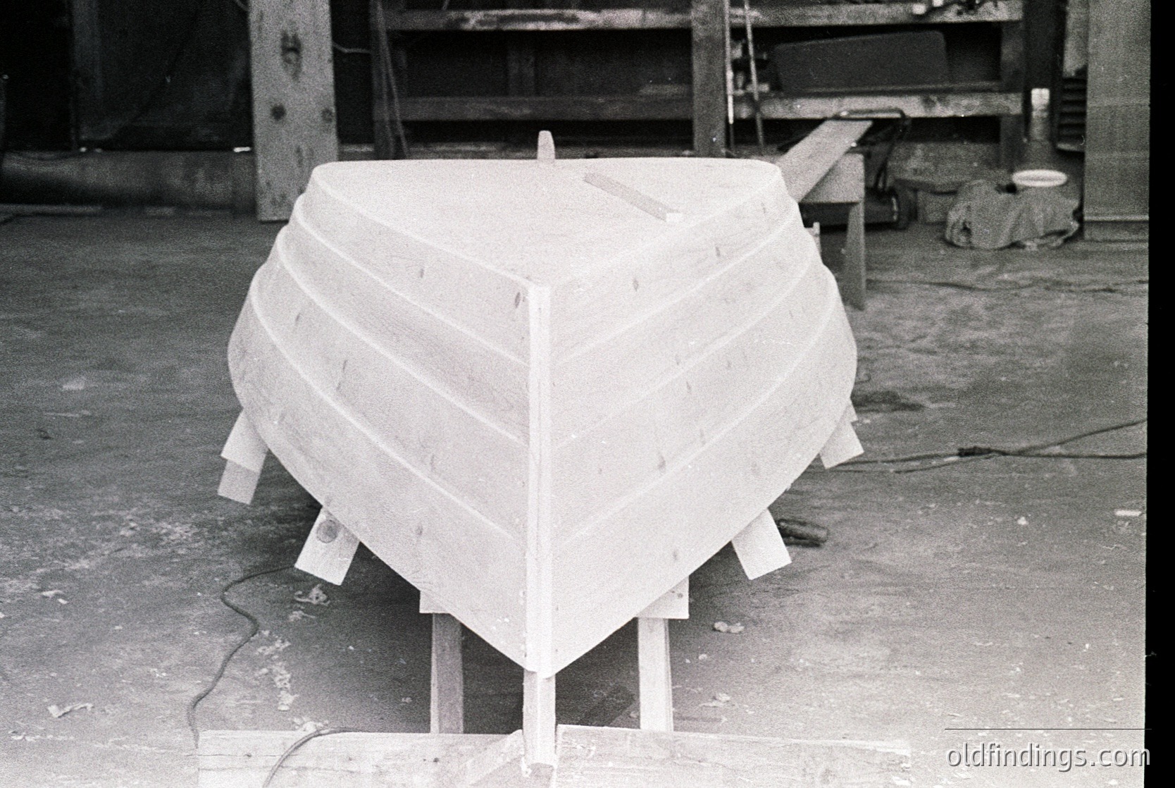 Unfinished wooden boat hull in a workshop, secured on wooden supports. Planking shows hand-cut seams, indicating traditional carpentry. Industrial setting with tools and machinery in background. Likely mid-20th century boatbuilding.