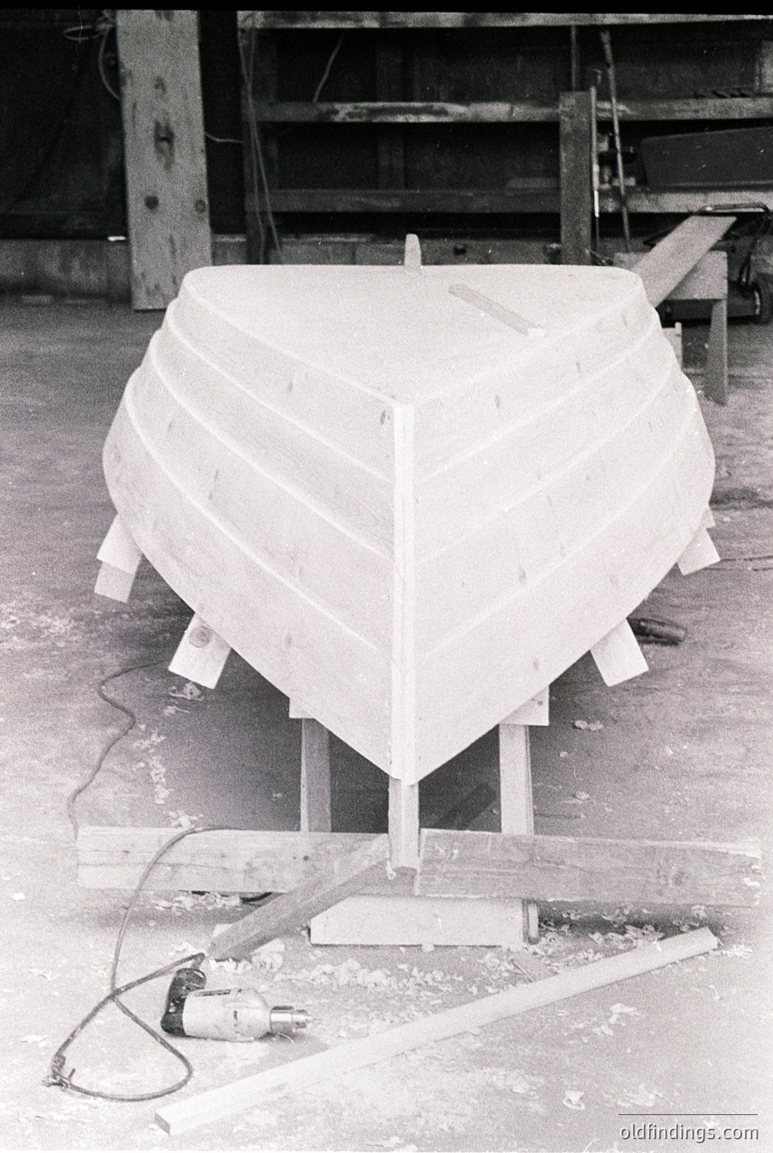 Mid-stage wooden boat hull construction in a workshop, featuring plank-on-bulkhead framing. Power tool and wooden supports visible beneath. Industrial setting with concrete floor and metal shelving in background.