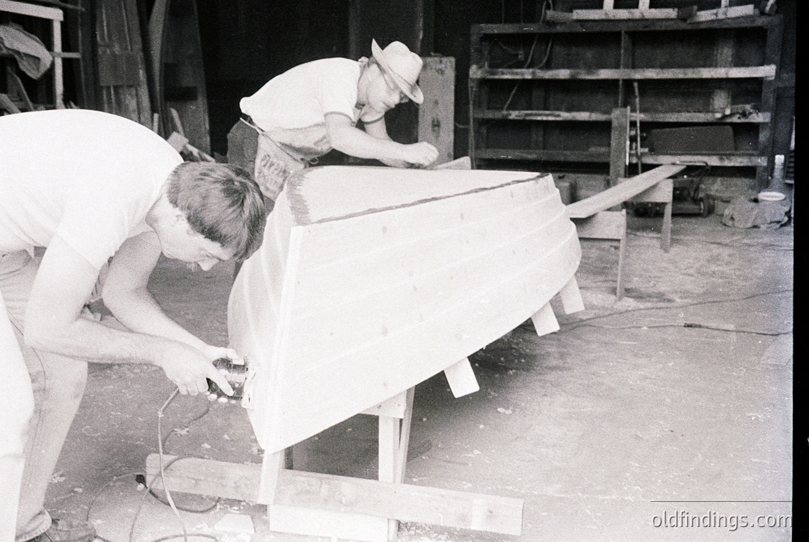Two men in 1950s-60s workwear sand and shape a large wooden boat hull in an industrial workshop. Visible tools include sandpaper, clamps, and a power sander. Shelving with wooden planks and tools in background.