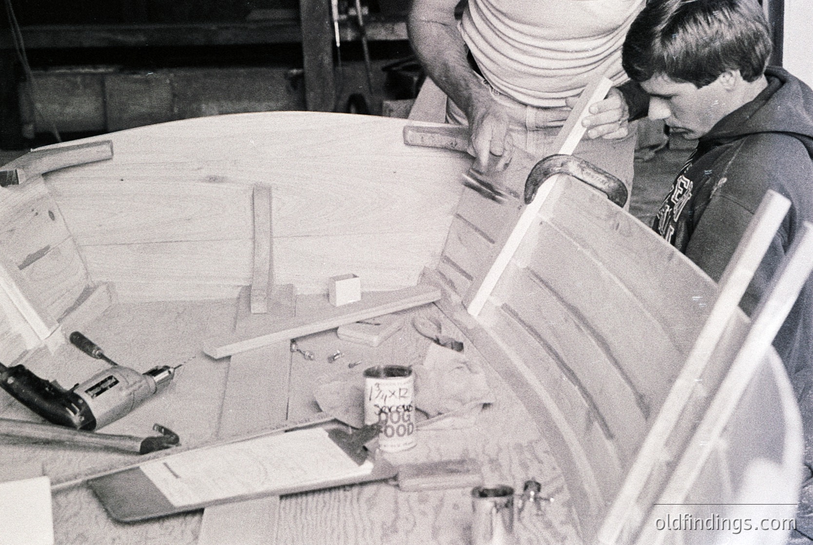 Two men collaborate on woodworking in a workshop, assembling a curved wooden frame. Tools like a hammer, clamps, and a measuring tape lie scattered on the workbench. Handwritten notes on a can suggest mid-20th century industrial or educational setting.