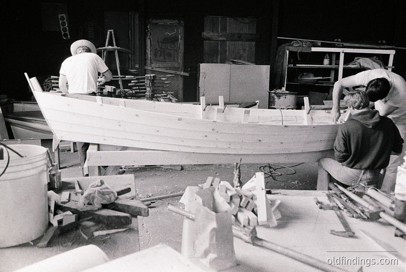Two craftsmen build a wooden boat in a workshop, surrounded by tools and materials. Mid-20th century industrial craftsmanship.