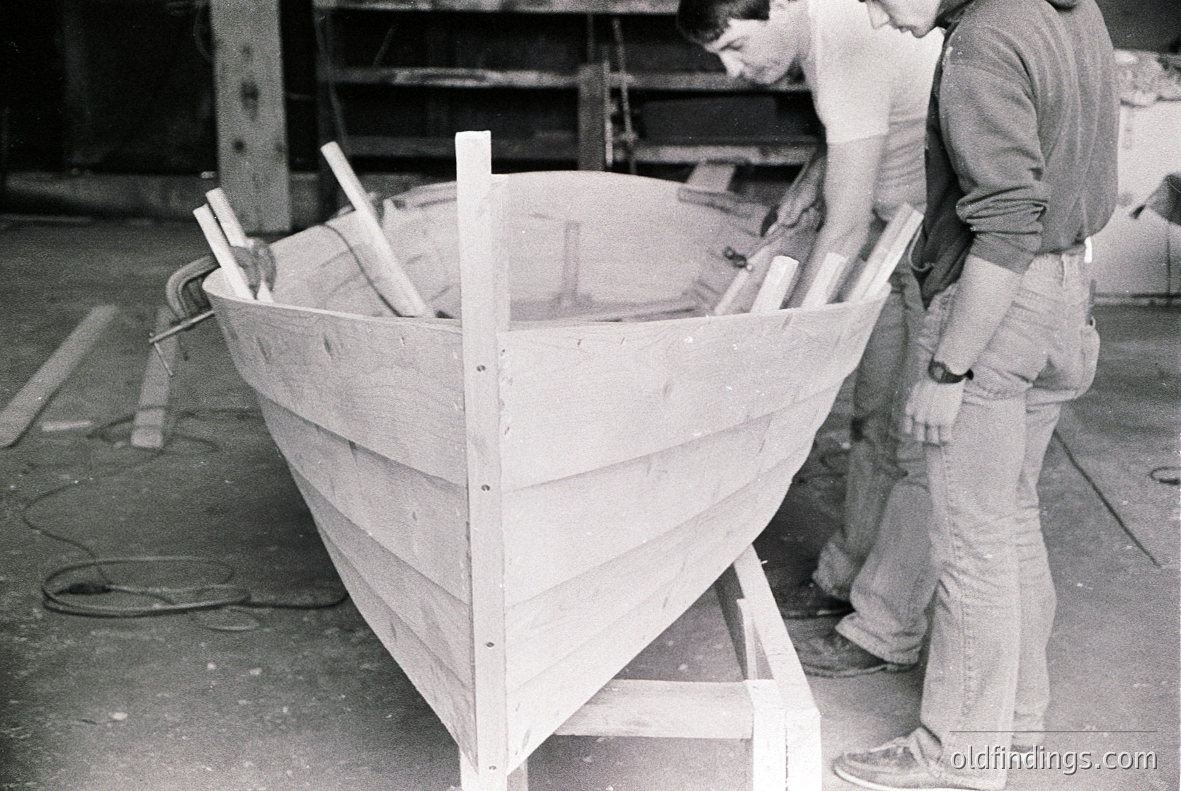 Handcrafted wooden boat under construction, likely mid-20th century. Two workers inspect planks and ribs in an industrial workshop setting. Raw materials and tools visible, suggesting traditional boatbuilding techniques. Potential maritime heritage.