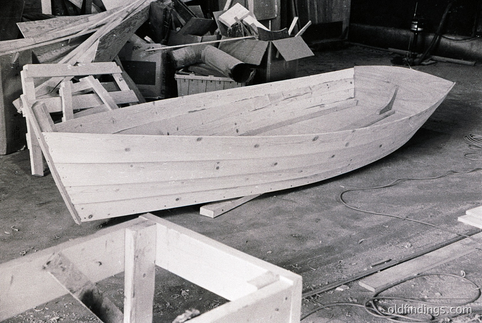 Unfinished wooden boat frame in a workshop, likely mid-construction. Ribbed hull with planks secured by nails, surrounded by lumber and tools. Industrial setting with concrete floor and metal pipes.