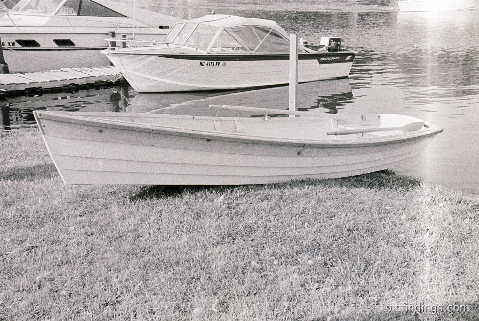 Classic wooden sailboat on grassy shore, mid-20th century design. Foreground boat features simple hull, transom stern, and minimal deck fittings. Background shows two motorboats with registration numbers. Likely a lakeside or marina setting, vintage .