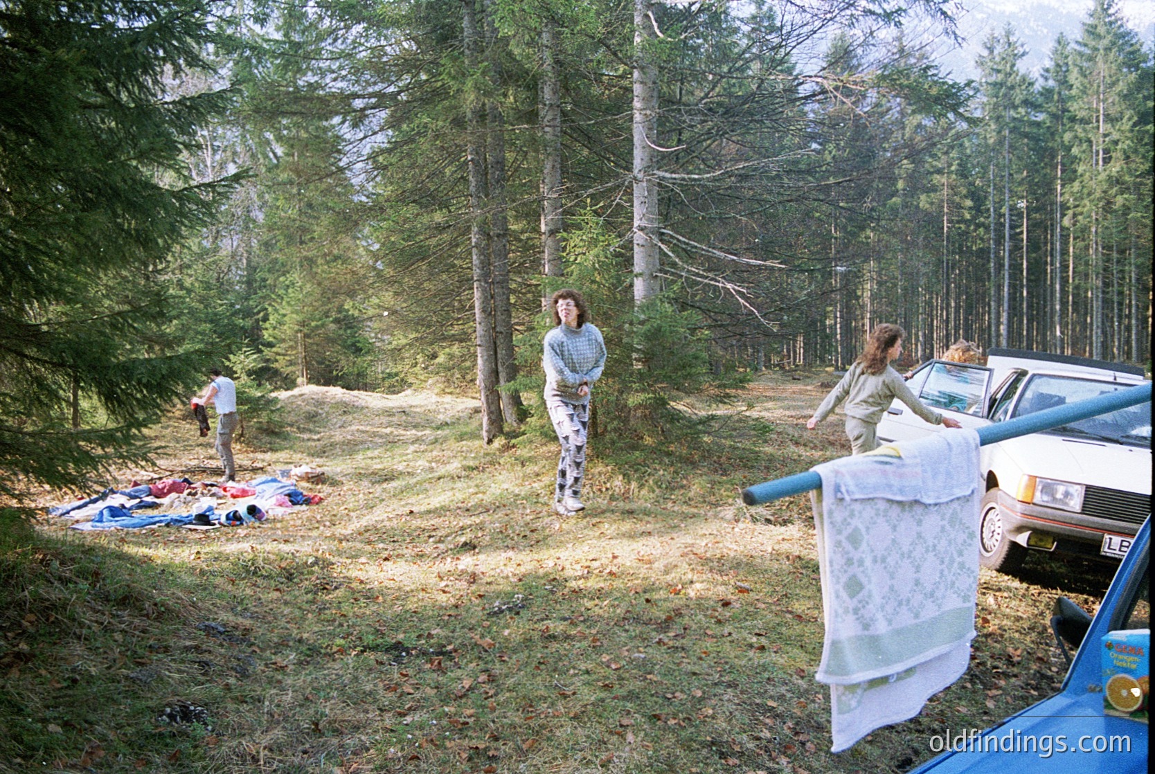 Forest picnic scene with vintage car: Three people setting up a spread on a dirt path surrounded by conifers. Spread includes blankets, a patterned cloth, and a white tablecloth. Car’s blue door visible on right. Likely 1970s–1990s outdoor leisure.