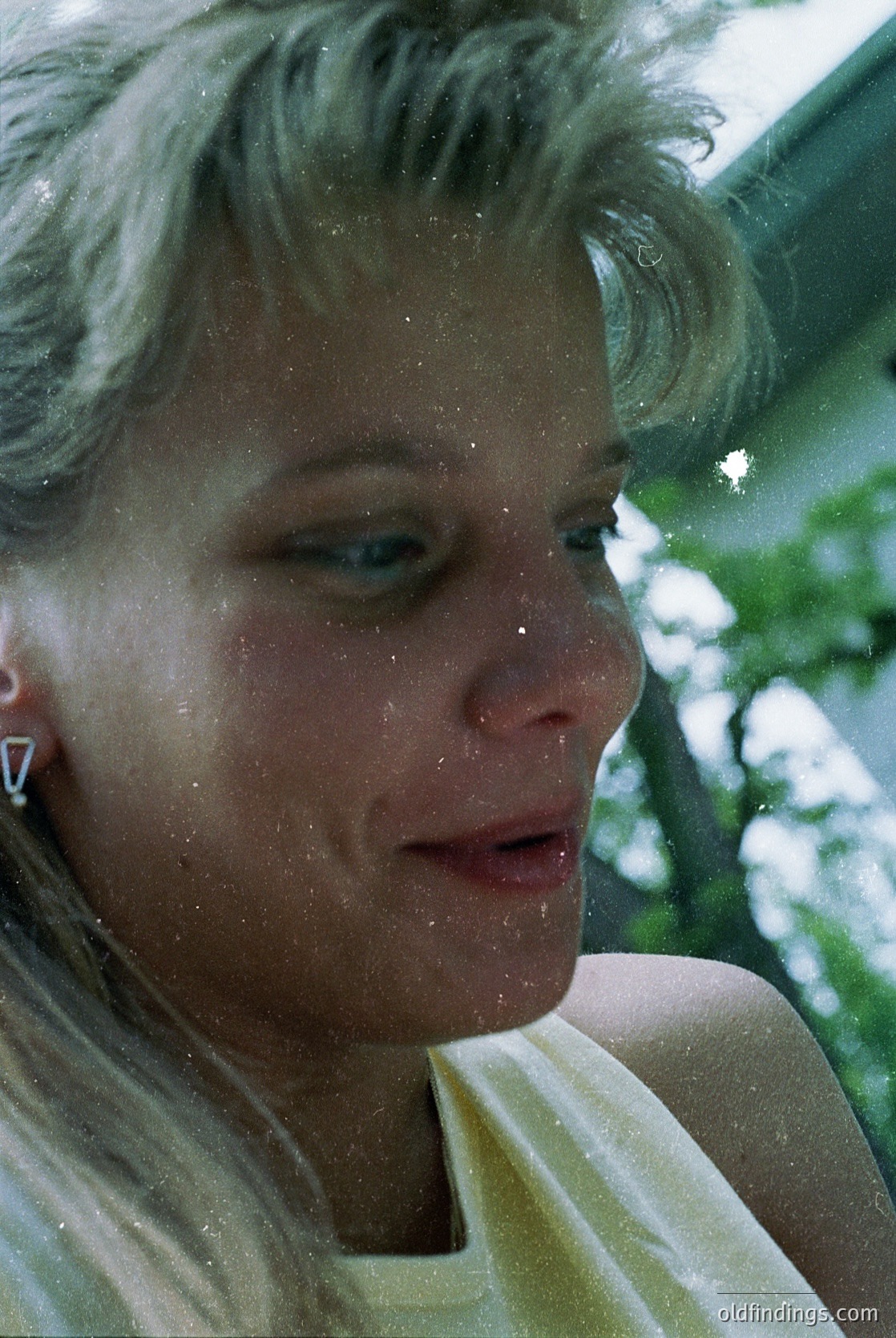 Close-up portrait of a woman with short, wavy blonde hair, captured mid-laugh or exhale. She wears minimalist white diamond stud earrings and a light-colored sleeveless top. Water droplets suggest a refreshing, outdoor setting—likely a poolside or seaside moment. Timeless, candid expression evokes joy and spontaneity.
