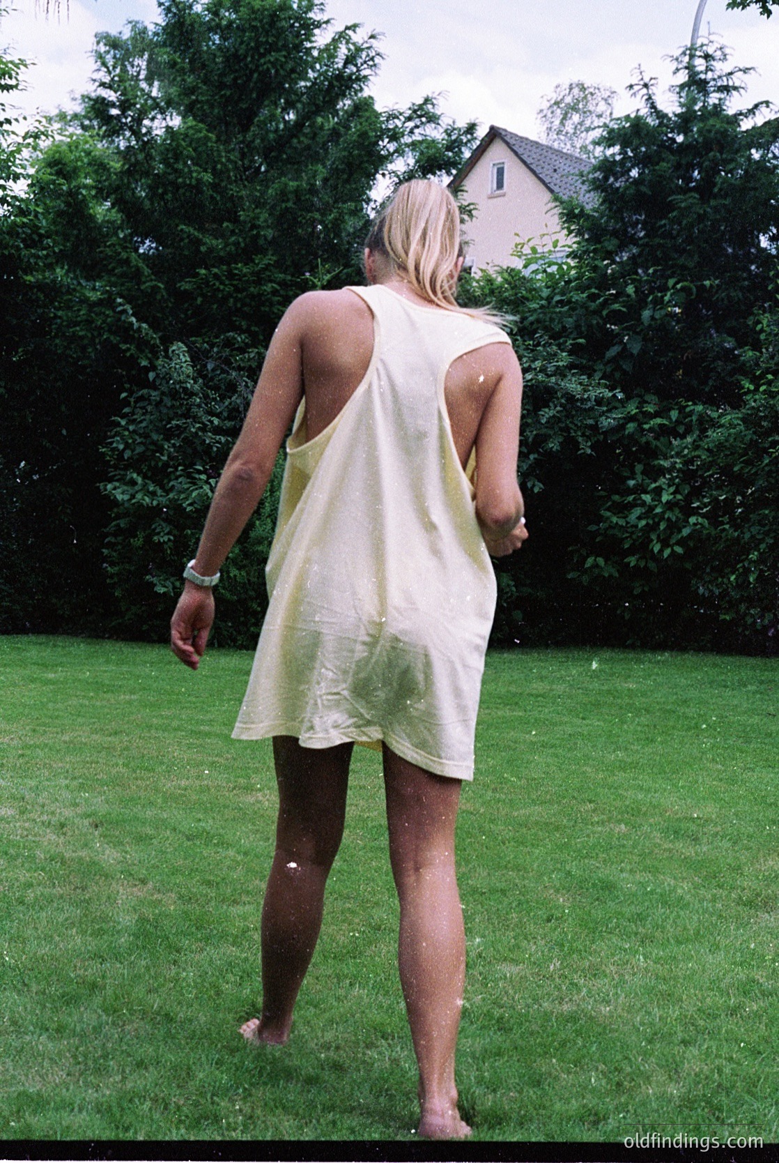 Backlit woman in a sleeveless, knee-length dress stands barefoot on lush green lawn, facing away. Subtle water droplets suggest recent rain or dew. Residential house with pitched roof and dense greenery in background. Mid-20th century suburban setting.